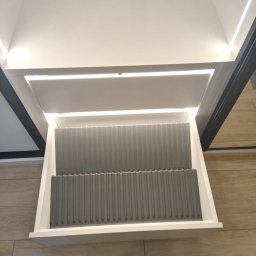 ROBERT DAWID - Wysuwana szafka z szufladami z szarymi organizerami na talerze i pokrywki, oświetlenie LED wewnątrz szafki, minimalistyczny design.