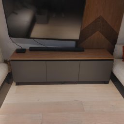 ROBERT DAWID - Szara szafka RTV z drewnianym blatem pod telewizorem, ustawiona między dwiema sofami w salonie z drewnianą ścianą dekoracyjną w tle.