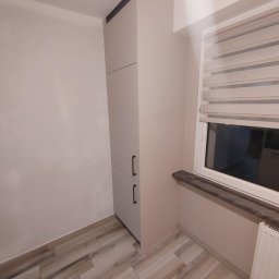 ROBERT DAWID - Wysoka, smukła szafa wnękowa w odcieniach beżu z czarnymi, minimalistycznymi uchwytami, zintegrowana ze ścianą w pomieszczeniu z drewnopodobną podłogą i oknem z żaluzjami.