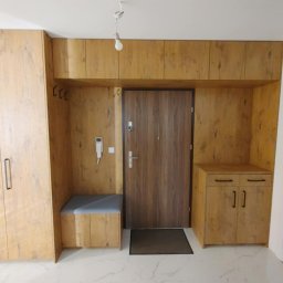 ROBERT DAWID - Przedpokój z zabudową meblową w odcieniach drewna, obejmującą szafę, siedzisko, szafki nad drzwiami i szafkę z szufladami, z widocznym domofonem i minimalistycznym oświetleniem.