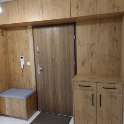 ROBERT DAWID - Przedpokój z zabudową meblową w odcieniach drewna, obejmującą szafki górne, siedzisko z szarą tapicerką, szafkę na buty z czarnymi uchwytami oraz drzwi wejściowe.