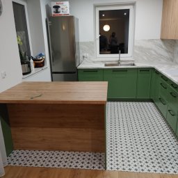 ROBERT DAWID - Jasna kuchnia z zielonymi szafkami, drewnianym blatem wyspowym i wzorzystą podłogą. W oknie widać odbicie osoby robiącej zdjęcie.