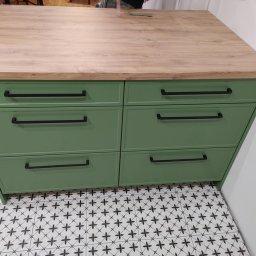 ROBERT DAWID - Zielona komoda z sześcioma szufladami i drewnianym blatem, czarne, poziome uchwyty, wzorzysta podłoga.