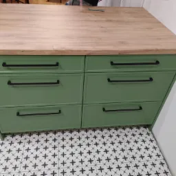 Zielona komoda z sześcioma szufladami i drewnianym blatem, czarne, poziome uchwyty, wzorzysta podłoga.