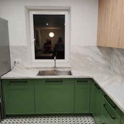 ROBERT DAWID - Nowoczesna kuchnia z zielonymi szafkami, marmurowym blatem i backsplashiem, stalowym zlewem pod oknem, i drewnianymi szafkami górnymi; odbicie osób w oknie.