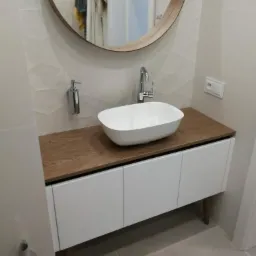 Nowoczesna łazienka z owalną umywalką nablatową na szafce z drewnianym blatem i białymi frontami, okrągłe lustro w drewnianej ramie, ściany z delikatną teksturą.