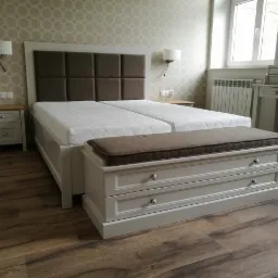 Jasnoszara sypialnia z łóżkiem kontynentalnym, tapicerowanym wezgłowiem i skrzynią z siedziskiem w tym samym odcieniu, na drewnianej podłodze.