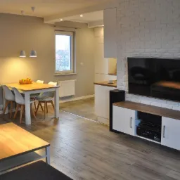 Jasne wnętrze salonu z jadalnią i aneksem kuchennym, drewniana podłoga, biała cegła na ścianie, stół z owocami, minimalistyczne lampy wiszące.