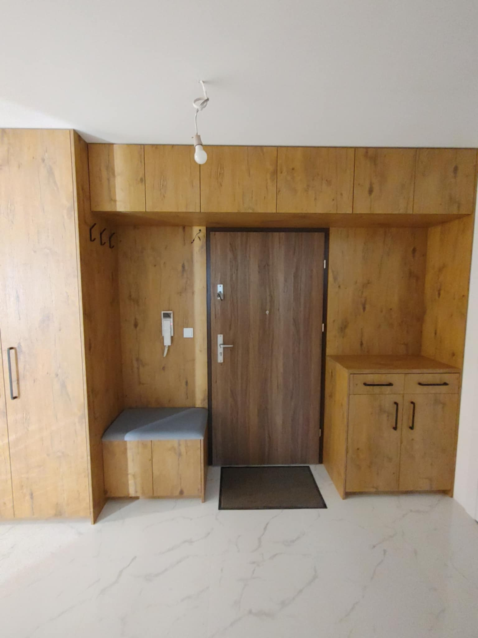 Przedpokój z zabudową meblową w odcieniach drewna, obejmującą szafę, siedzisko, szafki nad drzwiami i szafkę z szufladami, z widocznym domofonem i minimalistycznym oświetleniem.