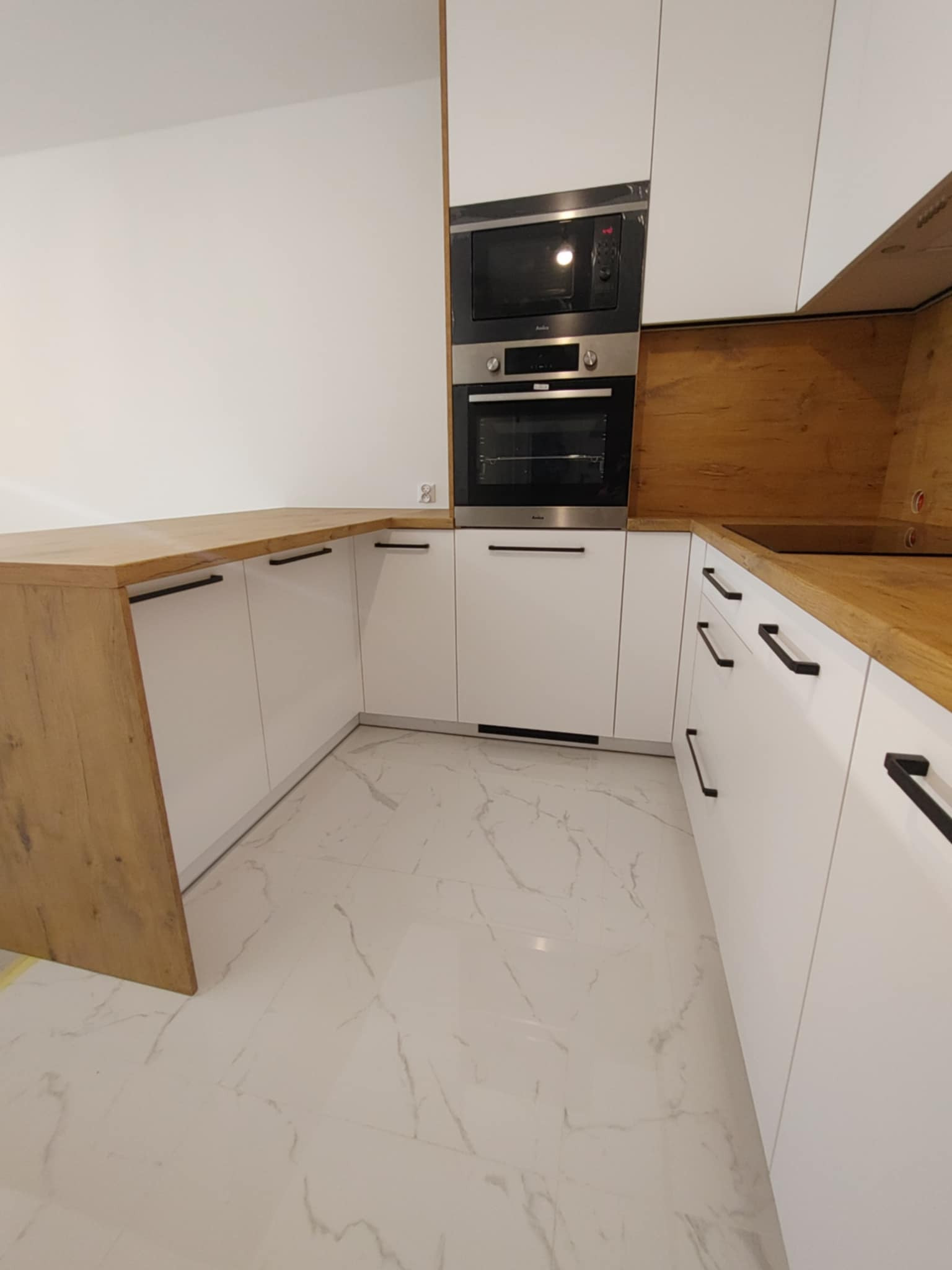 Nowoczesna kuchnia z białymi frontami i drewnianym blatem, wyposażona w piekarnik i mikrofalówkę w zabudowie, na podłodze białe płytki imitujące marmur.