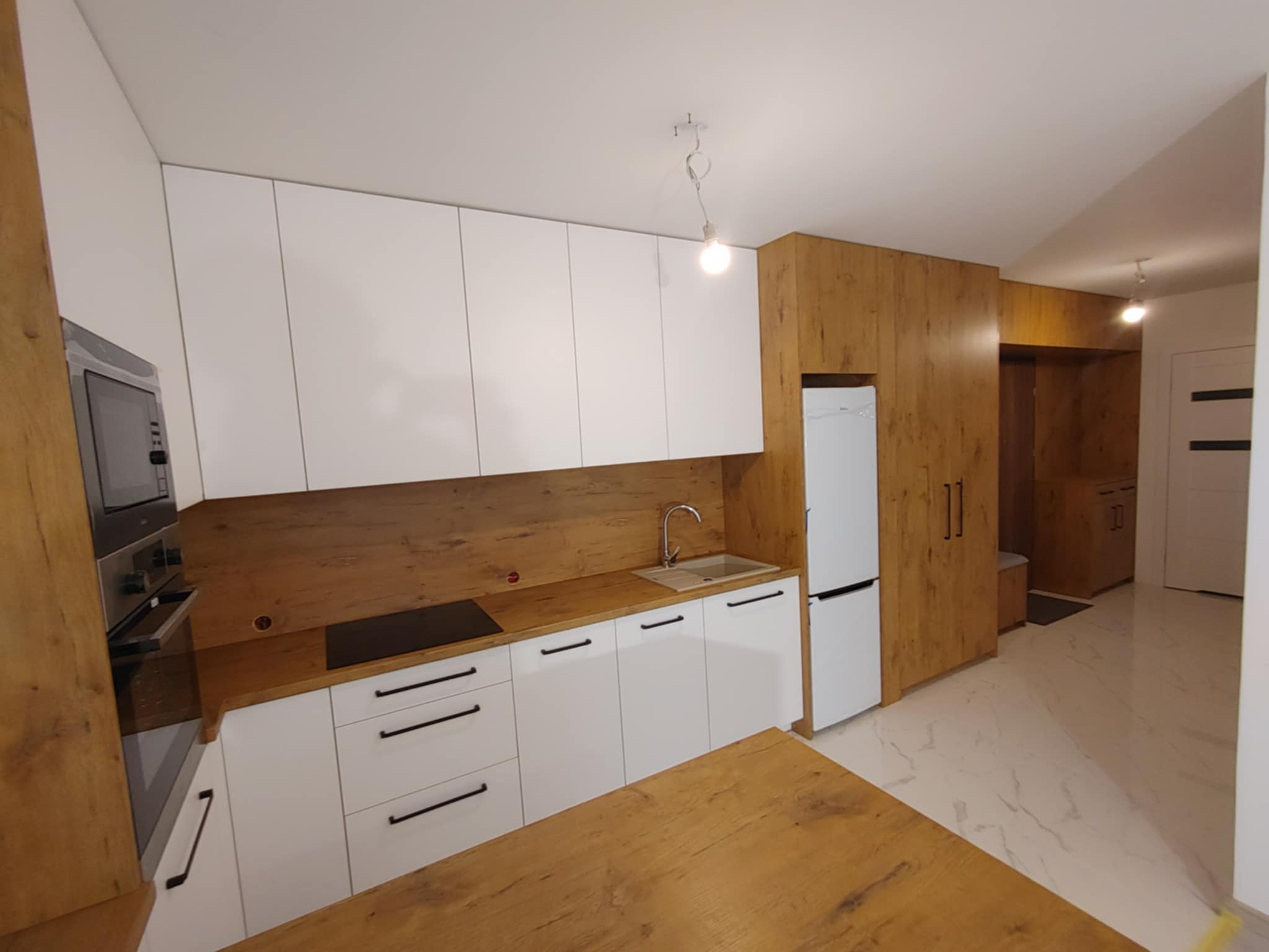 Nowoczesna kuchnia z białymi szafkami i drewnianymi akcentami, widok na blat kuchenny, zlew, płytę indukcyjną i lodówkę, całość dopełniona drewnianą zabudową ścienną.