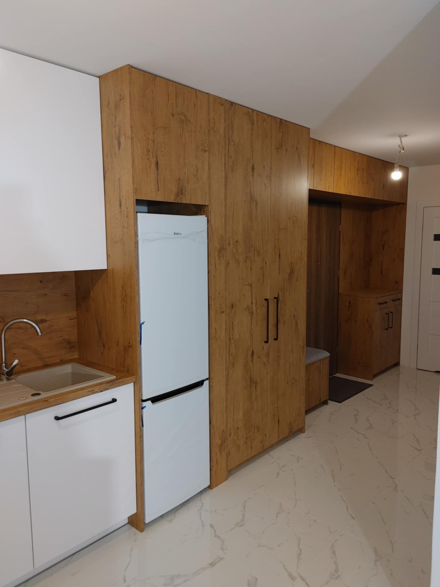 Nowoczesna zabudowa kuchenna z frontami w kolorze drewna i białymi szafkami, zintegrowana z szafą wnękową i miejscem do siedzenia przy wejściu, na marmurowej podłodze.