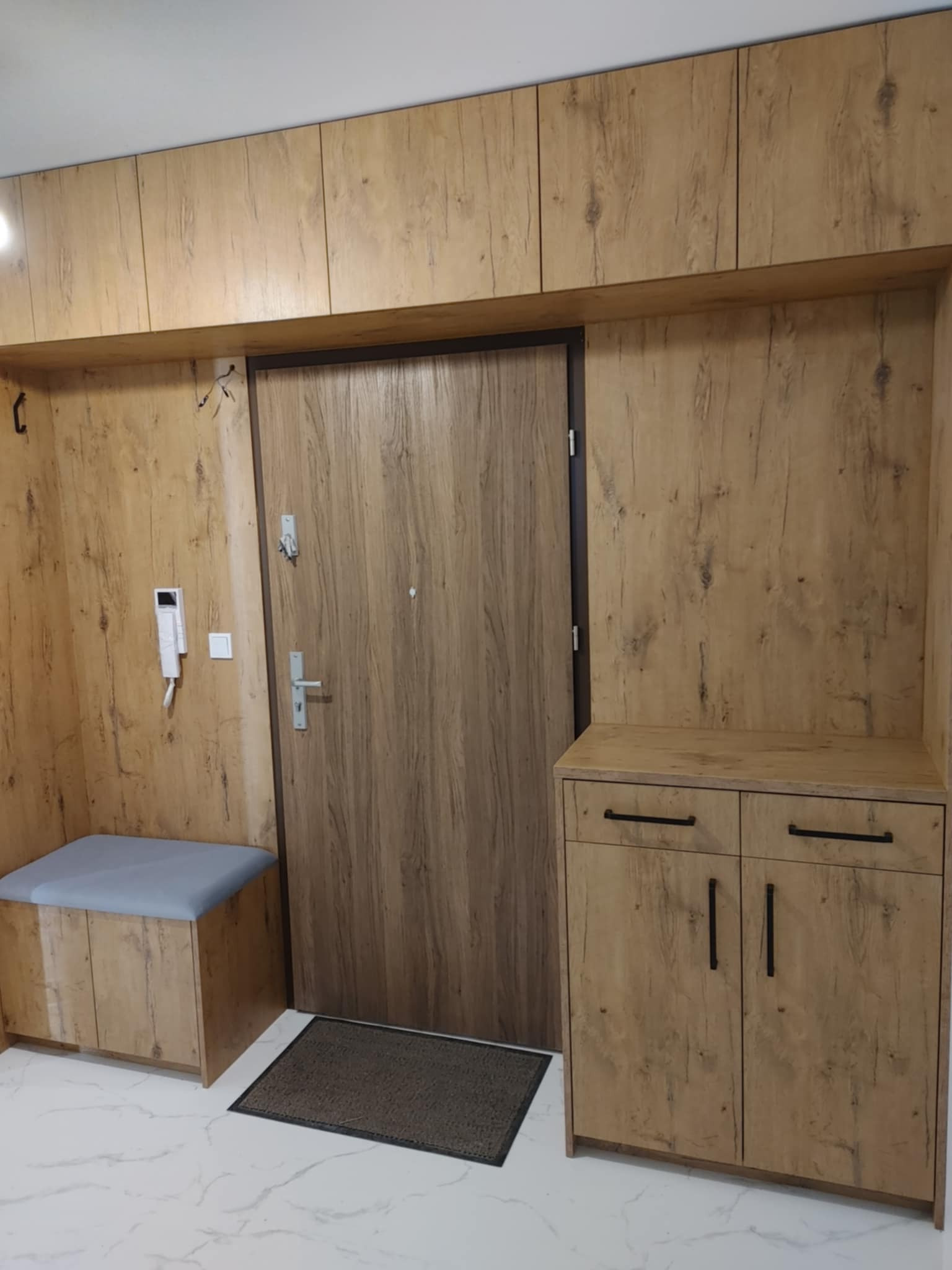 Przedpokój z zabudową meblową w odcieniach drewna, obejmującą szafki górne, siedzisko z szarą tapicerką, szafkę na buty z czarnymi uchwytami oraz drzwi wejściowe.