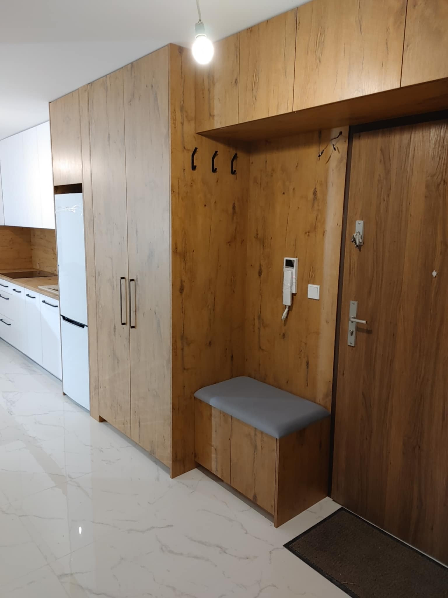 Przedpokój z drewnianą zabudową obejmującą szafę, siedzisko z wieszakami i drzwi wejściowe, zintegrowany z aneksem kuchennym z białymi szafkami.