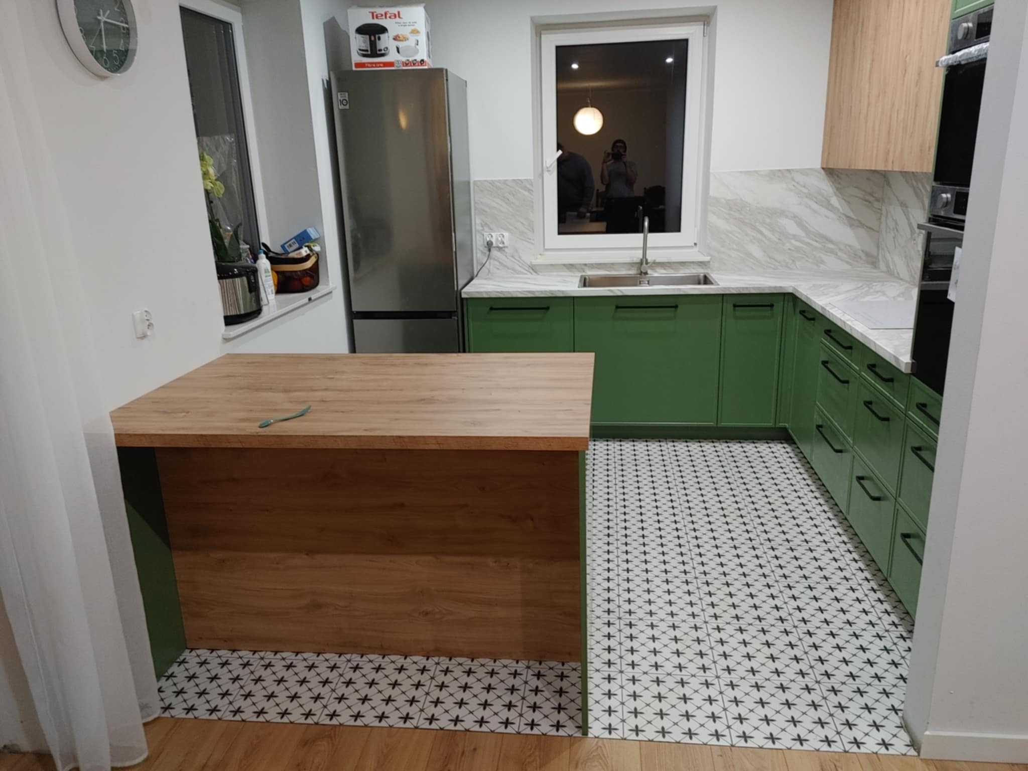 Jasna kuchnia z zielonymi szafkami, drewnianym blatem wyspowym i wzorzystą podłogą. W oknie widać odbicie osoby robiącej zdjęcie.