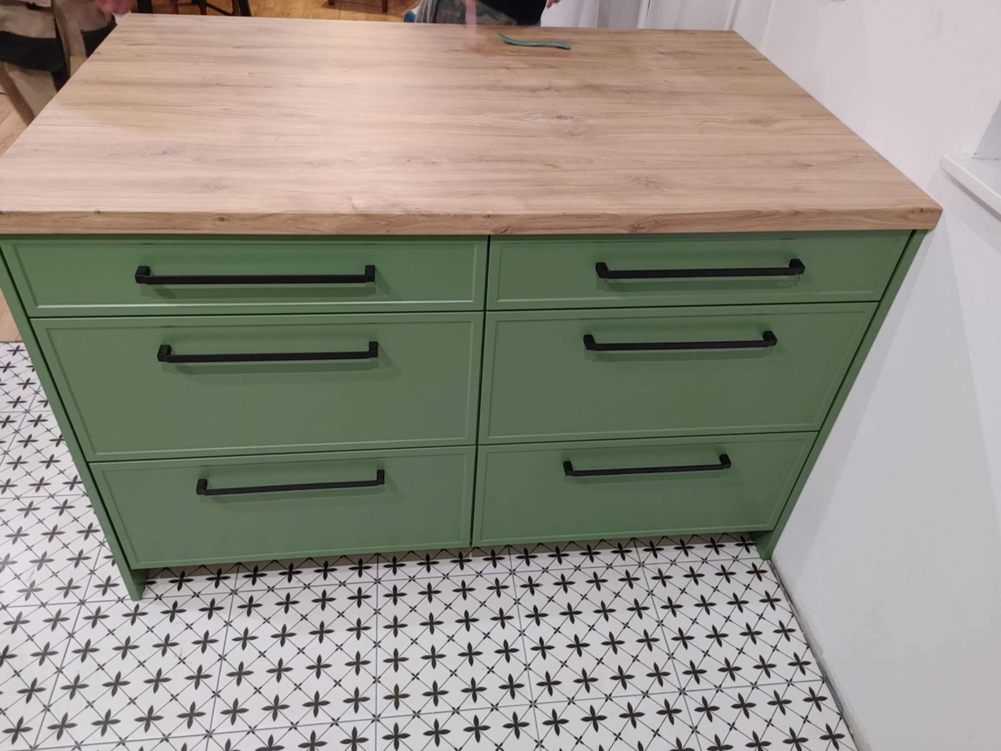 Zielona komoda z sześcioma szufladami i drewnianym blatem, czarne, poziome uchwyty, wzorzysta podłoga.