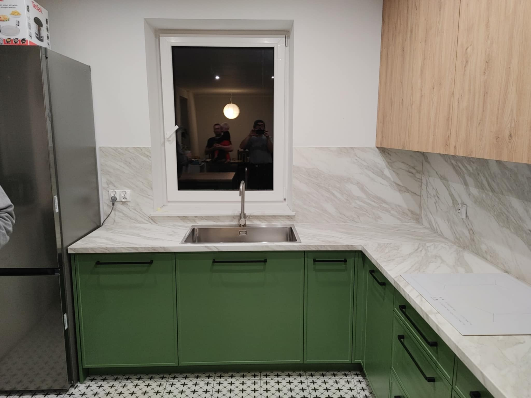 Nowoczesna kuchnia z zielonymi szafkami, marmurowym blatem i backsplashiem, stalowym zlewem pod oknem, i drewnianymi szafkami górnymi; odbicie osób w oknie.