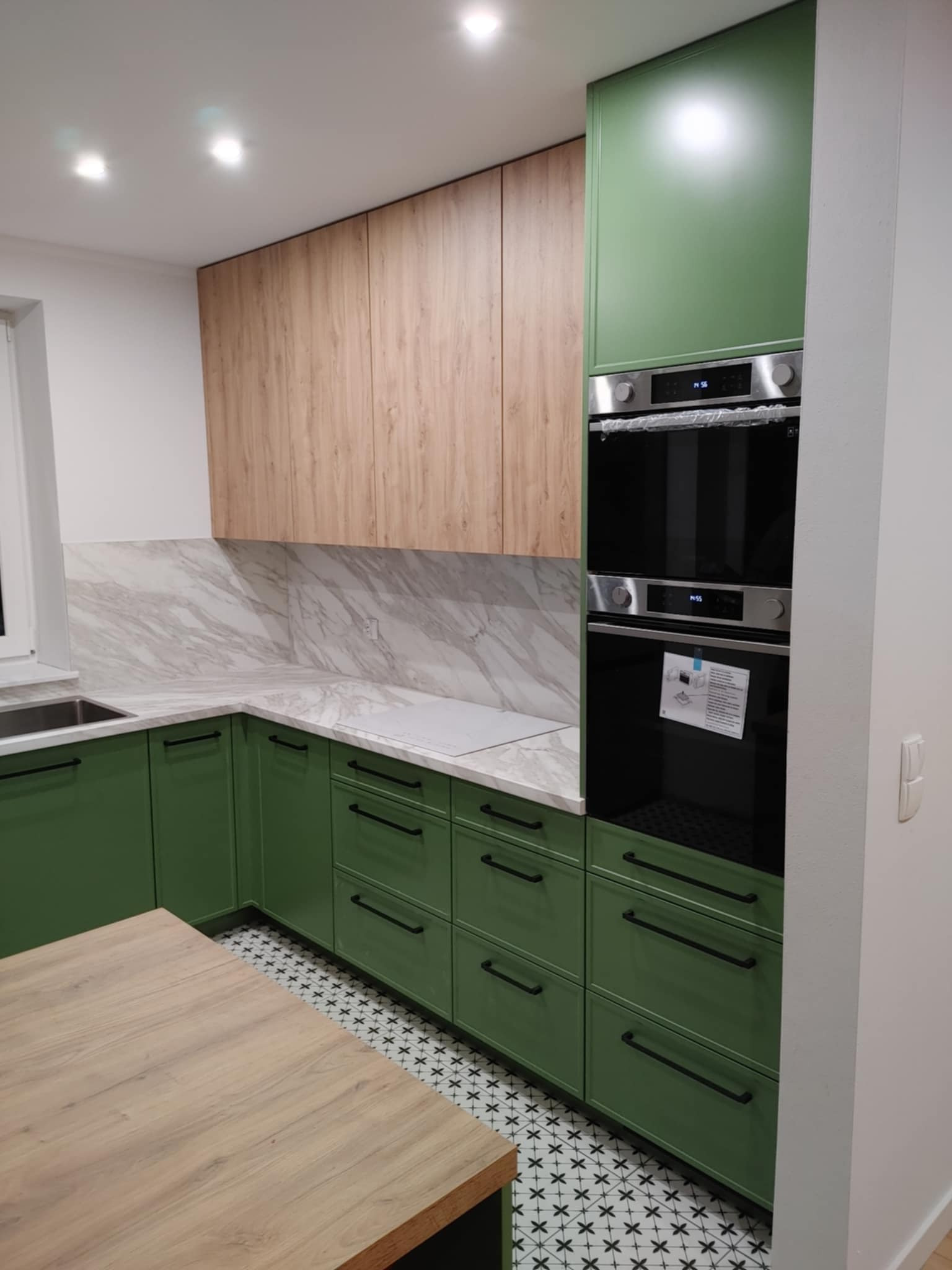 Nowoczesna kuchnia z zielonymi szafkami, drewnianymi górnymi szafkami i marmurowym blatem, z wbudowanym piekarnikiem i płytą indukcyjną.