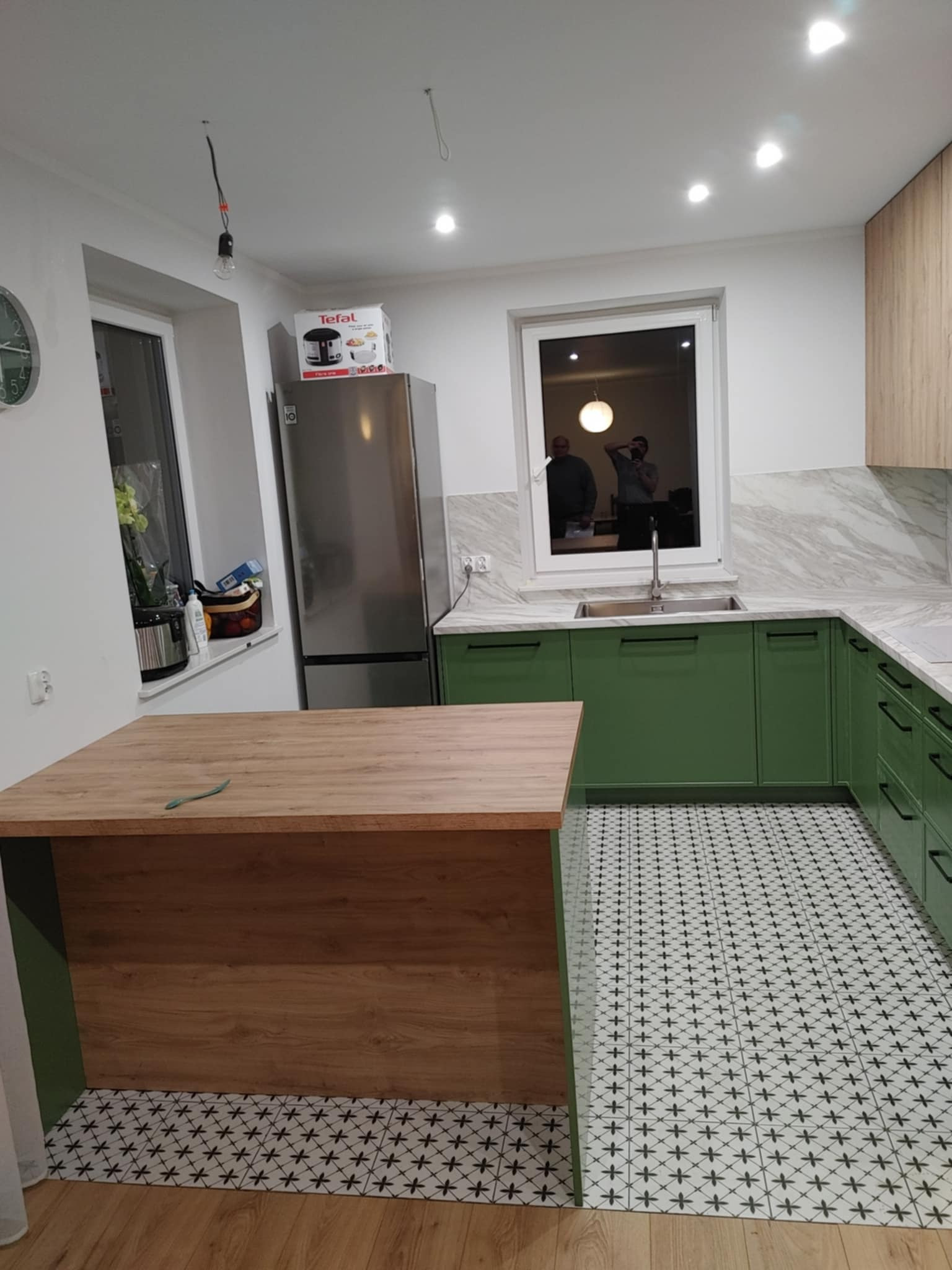 Nowoczesna kuchnia z zielonymi szafkami, blatem imitującym marmur i wyspą z drewnianym blatem, wzorzysta podłoga, widoczne odbicie dwóch osób w oknie.