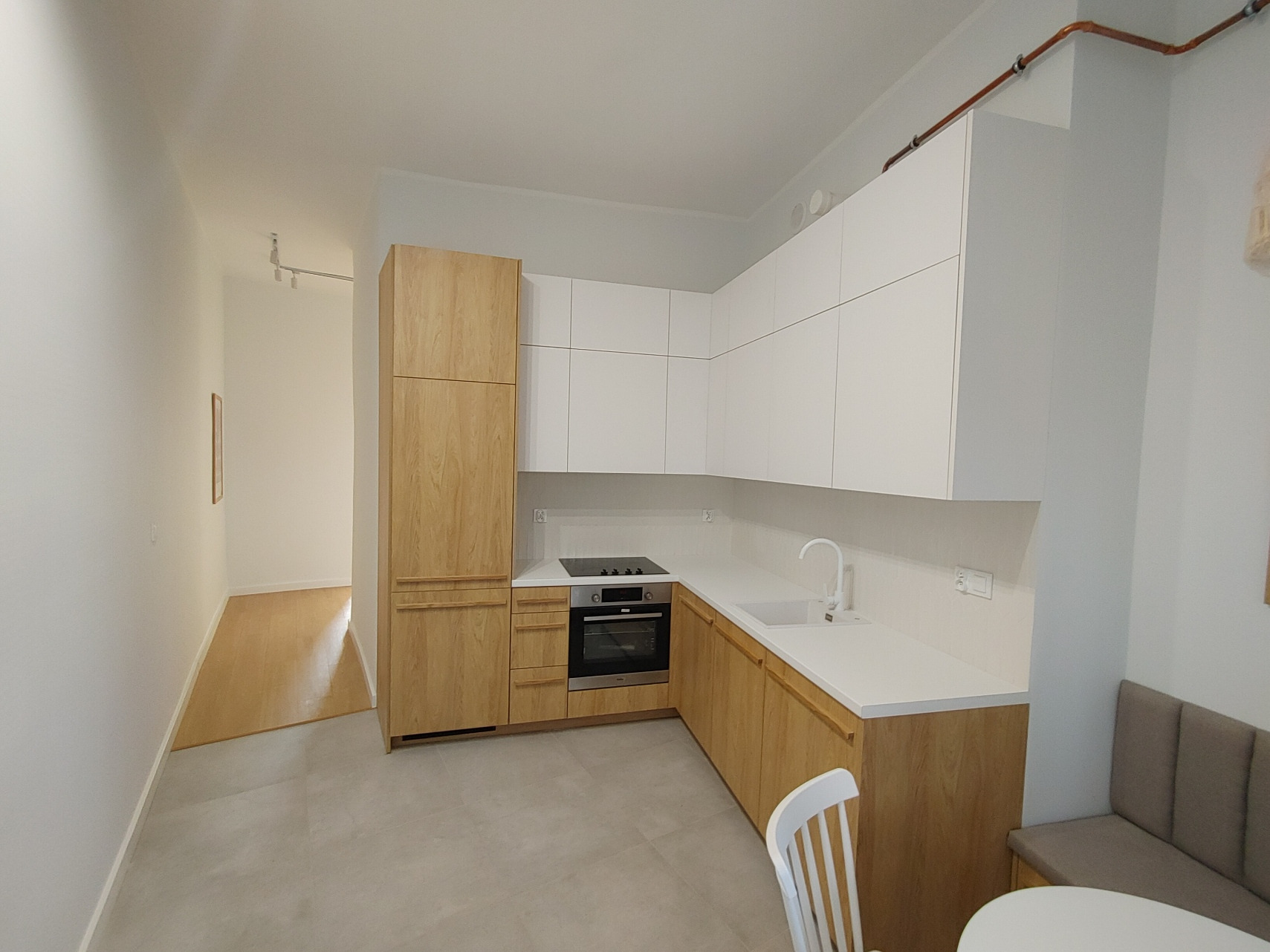 Jasna kuchnia z drewnianymi frontami i białymi szafkami wiszącymi, minimalistyczny design, widoczne rury miedziane pod sufitem, krzesło i stół w części jadalnej.