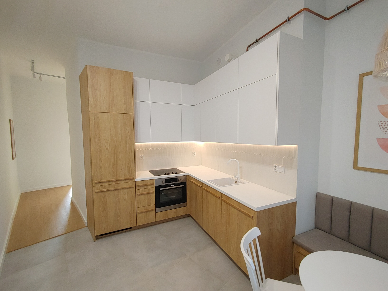 Jasna kuchnia z zabudową na wymiar: drewniane fronty dolne i wysoka szafa, białe szafki górne, blat z białego konglomeratu, oświetlenie LED podszafkowe, widoczne miedziane rury na suficie.