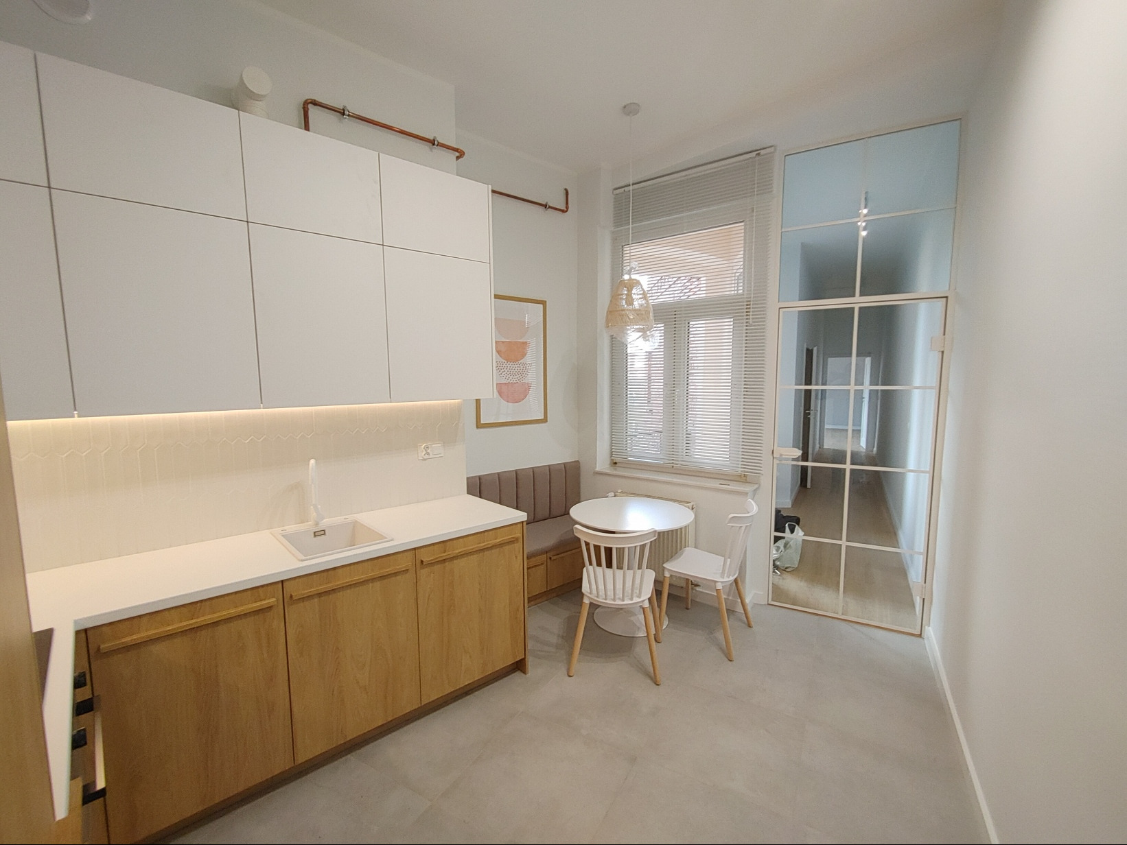 Nowoczesna, minimalistyczna kuchnia z drewnianymi frontami szafek dolnych i białymi szafkami wiszącymi, oświetlona listwą LED pod szafkami, z okrągłym stołem i krzesłami w rogu, oraz szklanymi...