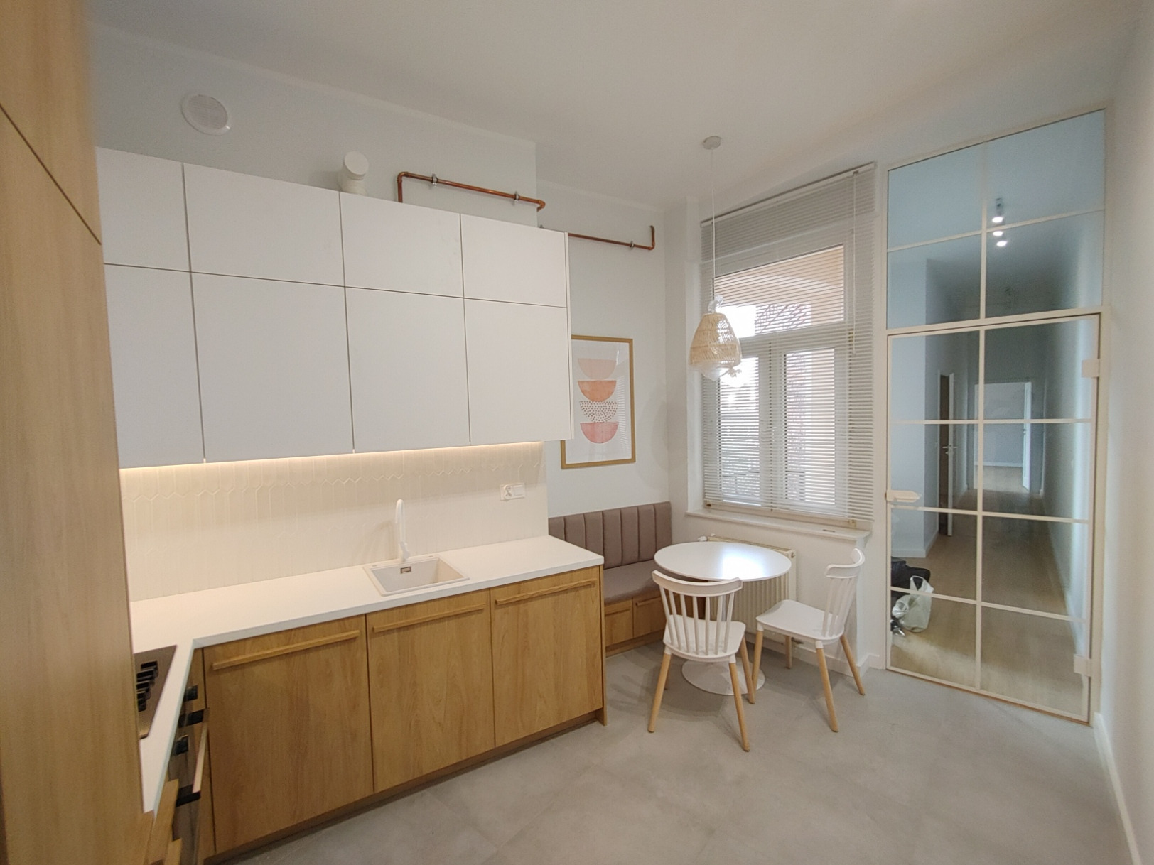 Jasna, nowoczesna kuchnia z białymi szafkami górnymi i drewnianymi dolnymi, okrągły stół z krzesłami przy oknie, szklane drzwi w stylu industrialnym, dekoracyjny panel nad blatem.