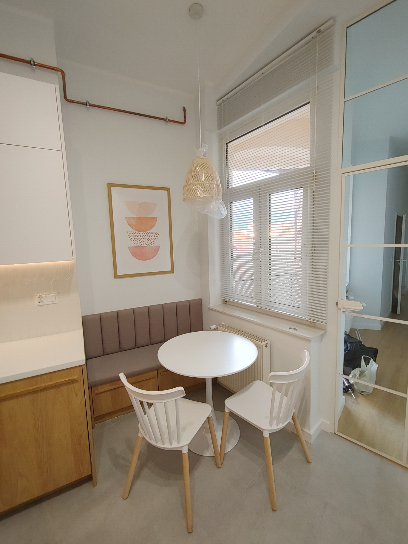 Jasna kuchnia z narożną zabudową i okrągłym stołem z krzesłami, nad stołem wisi lampa z abażurem w stylu boho, na ścianie grafika w ramie.