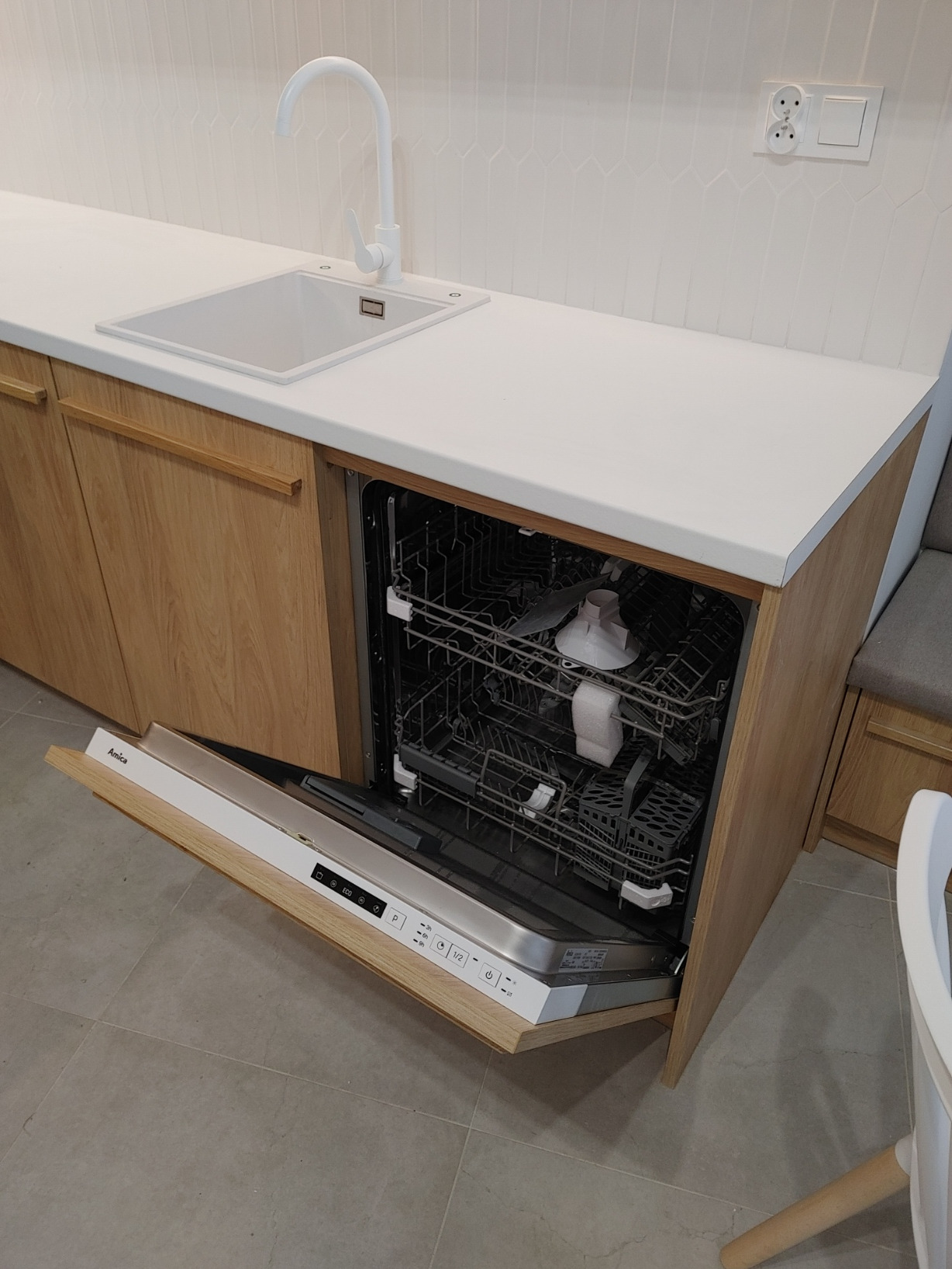 Nowoczesna kuchnia z drewnianymi frontami, białym blatem i zlewozmywakiem, z otwartą zmywarką Amica, ukazująca wnętrze i umieszczone w niej naczynia.