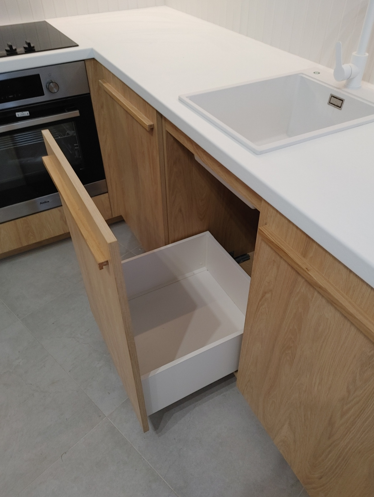 Fragment zabudowy kuchennej z jasnego drewna, otwarty biały szuflada pod blatem, zlew z białą baterią, płyta grzewcza i piekarnik w tle.