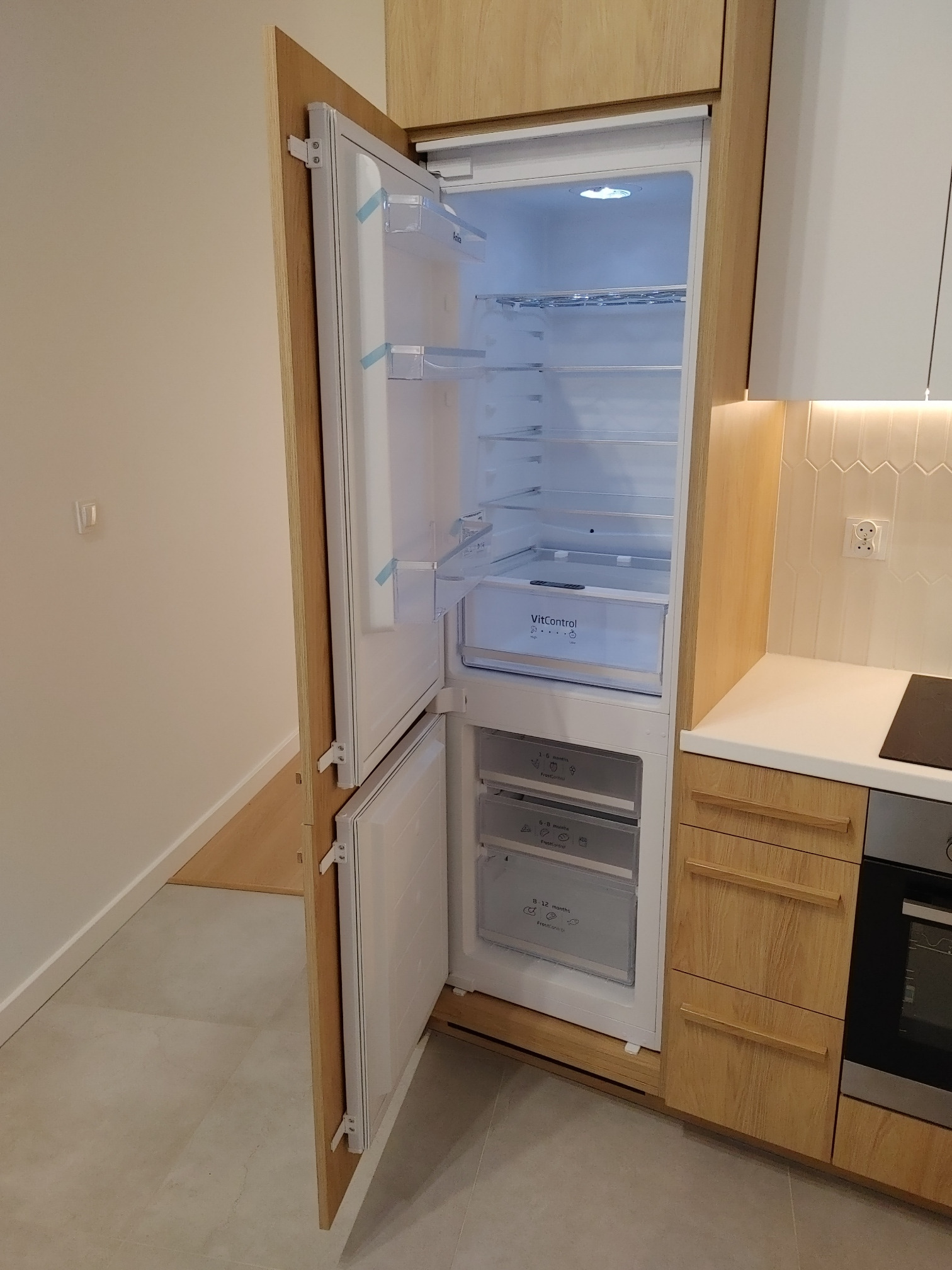 Otwarta lodówka w zabudowie kuchennej z drewnianymi frontami, widoczne półki i szuflady wewnątrz, oświetlenie LED, minimalistyczny styl.