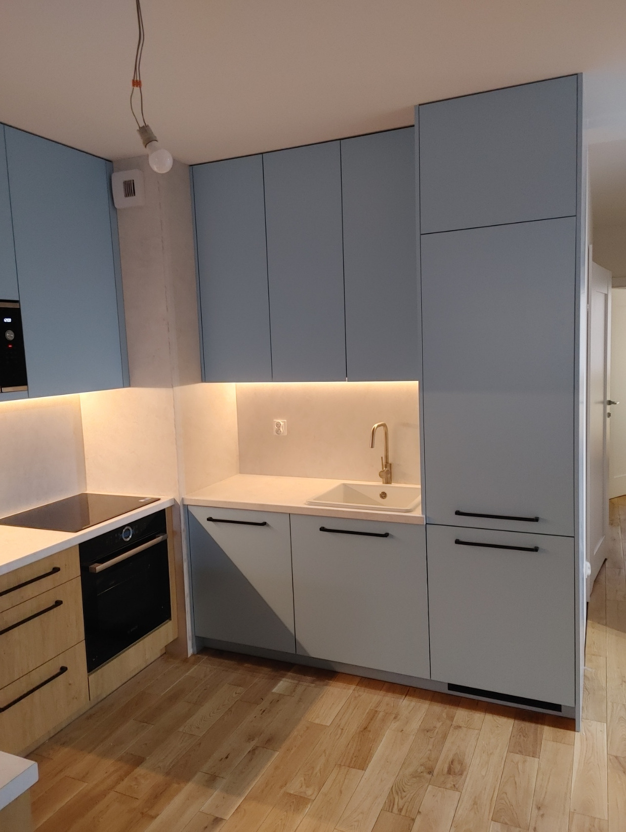 Nowoczesna kuchnia z jasnoniebieskimi szafkami, drewnianymi akcentami i złotą armaturą, oświetlona taśmą LED pod szafkami wiszącymi, z widoczną płytą indukcyjną i piekarnikiem.