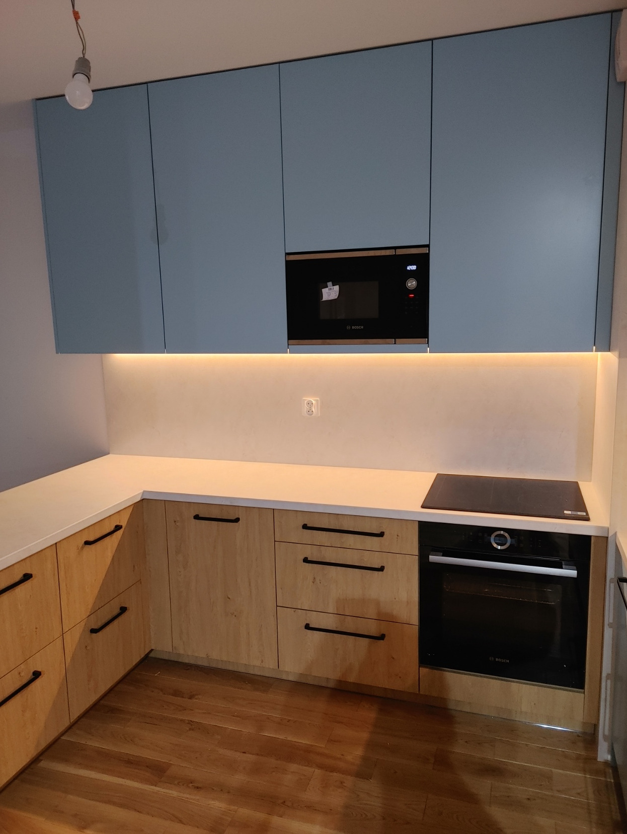 Nowoczesna kuchnia z błękitnymi górnymi szafkami i drewnopodobnymi dolnymi, oświetleniem LED pod szafkami górnymi, wbudowanym piekarnikiem i mikrofalówką marki Bosch.