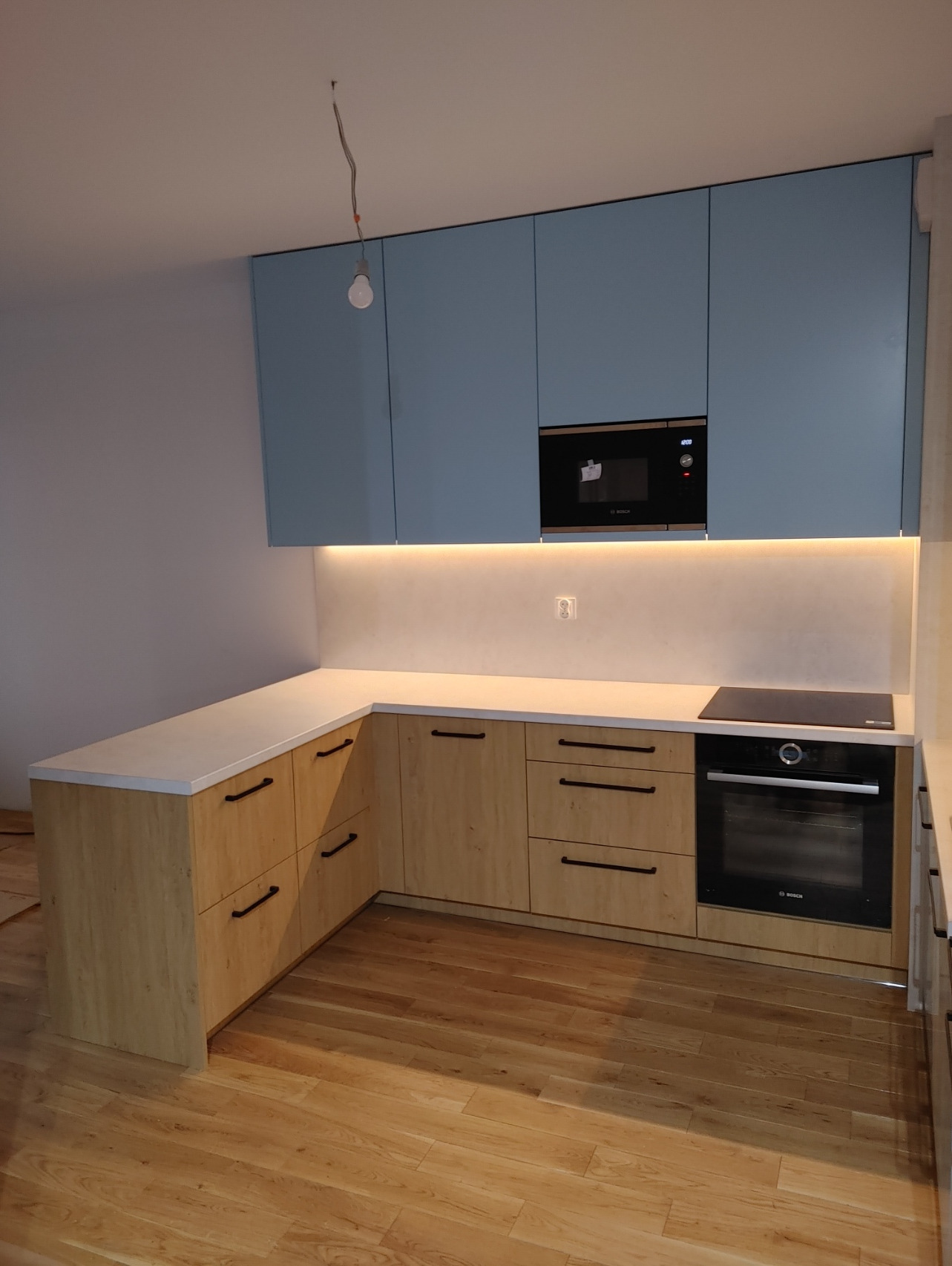 Nowoczesna kuchnia z jasnymi, drewnopodobnymi frontami szafek dolnych i błękitnymi szafkami górnymi, oświetleniem LED pod szafkami, czarnym piekarnikiem i płytą indukcyjną.