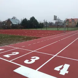 Czerwona bieżnia lekkoatletyczna z białymi numerami startowymi 1, 2, 3, 4, obok boisko do koszykówki i siatkówki, otoczone siatką, widok na zabudowania w tle, pochmurny dzień.