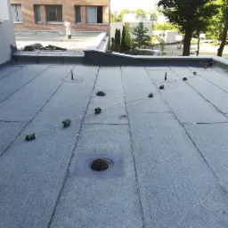 Płaski dach pokryty papą termozgrzewalną z widocznymi elementami instalacji odgromowej i wywietrznikami, budynek w tle.