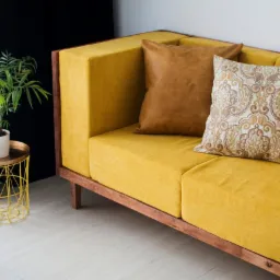 Żółta sofa z drewnianą ramą, brązową skórzaną poduszką i poduszką w ornamenty, obok stolik z rośliną doniczkową.