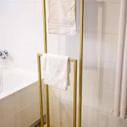 Nowoczesny, złoty, metalowy stojak na ręczniki z dwoma białymi ręcznikami w łazience z białymi płytkami i wanną.