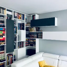 Biała, modułowa biblioteka na wymiar z ciemnoszarym akcentem, wypełniona książkami i dekoracjami, dopasowana do narożnika pokoju z białą sofą i żółtą poduszką.