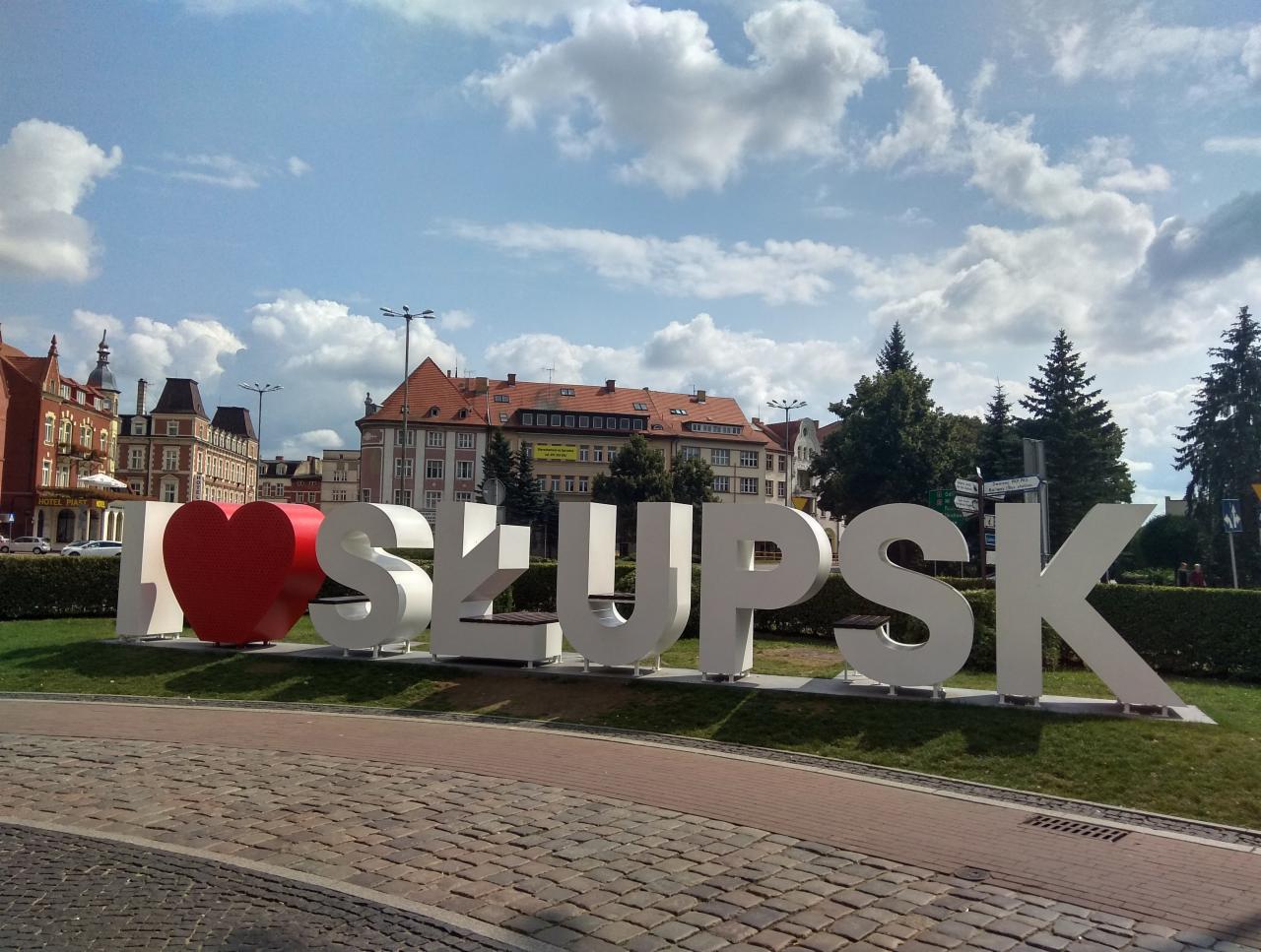 Instalacja artystyczna z napisem 'I LOVE SŁUPSK' na tle zabytkowej architektury miejskiej i błękitnego nieba z chmurami.