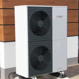 Biała pompa ciepła Vaillant aroTHERM plus zamontowana na betonowych podstawach obok brązowej elewacji z desek.