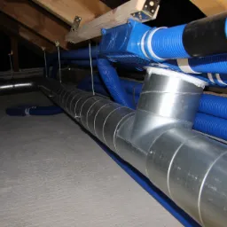 System wentylacyjny z rurami z blachy ocynkowanej i niebieskimi rurami karbowanymi, zamontowany na poddaszu z widoczną drewnianą konstrukcją dachu.