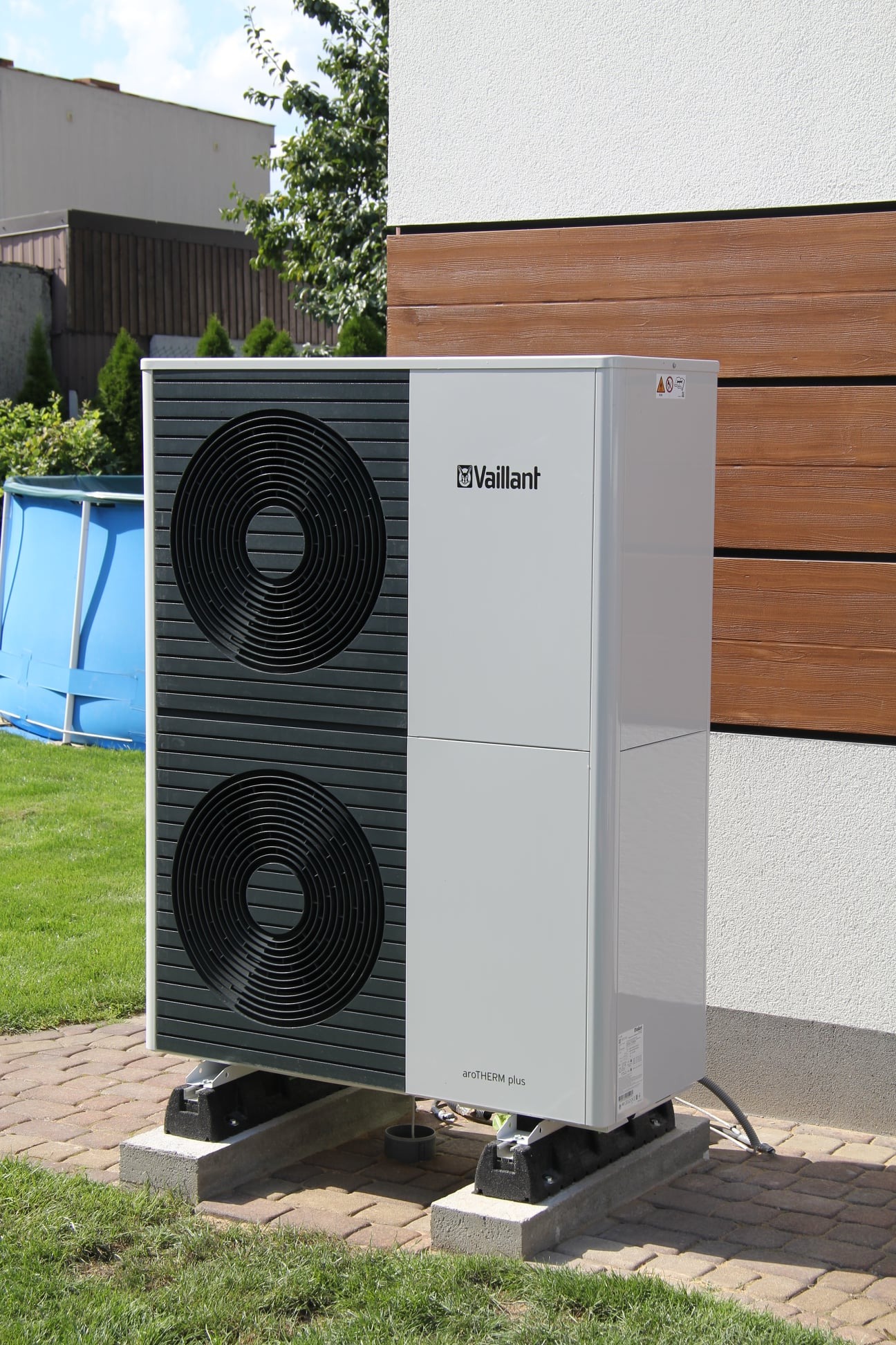 Zewnętrzna jednostka pompy ciepła Vaillant aroTHERM plus, zamontowana na betonowych podstawach przy ścianie budynku z drewnianą okładziną, widoczny trawnik i fragment basenu w tle.