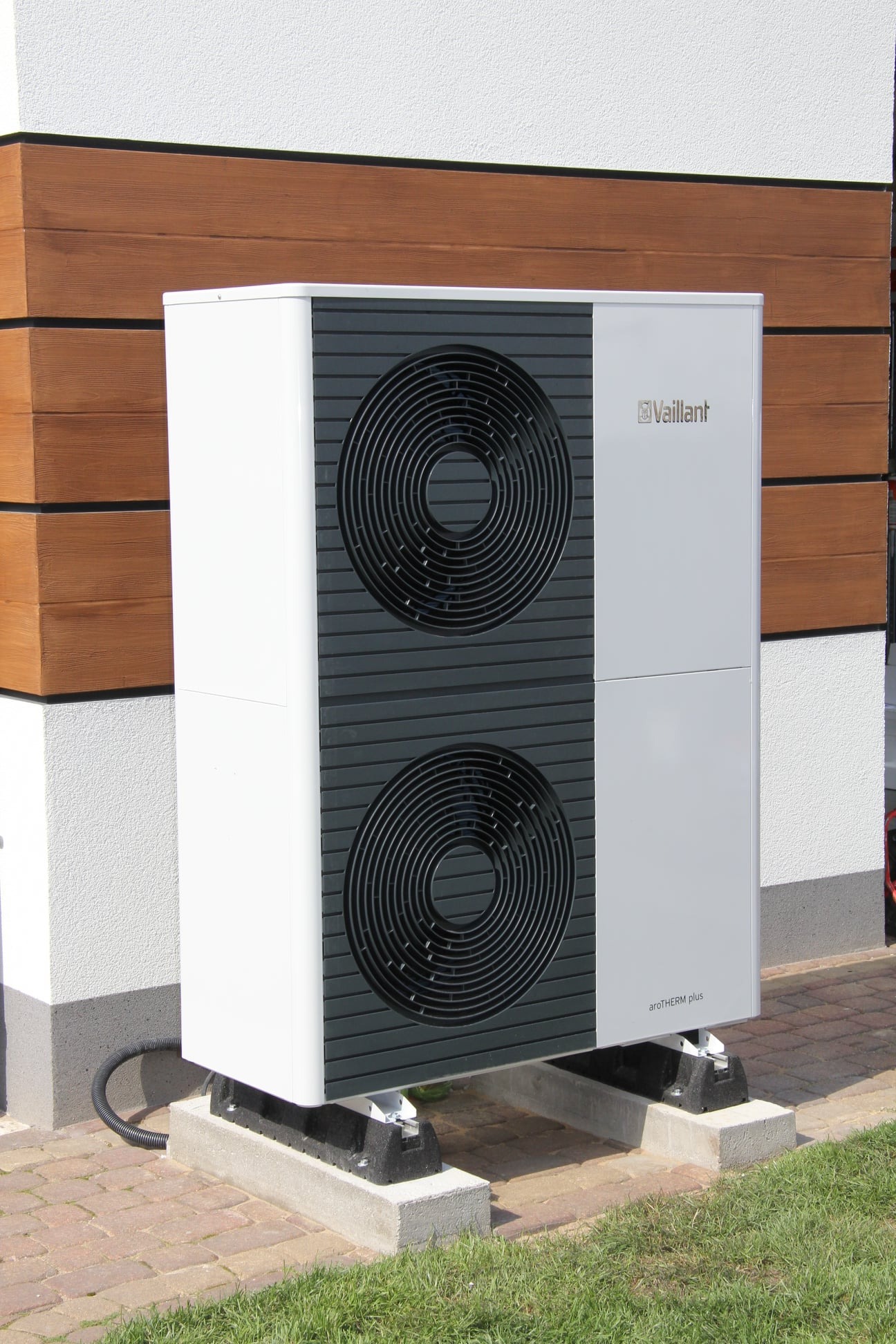 Biała pompa ciepła Vaillant aroTHERM plus zamontowana na betonowych podstawach obok brązowej elewacji z desek.