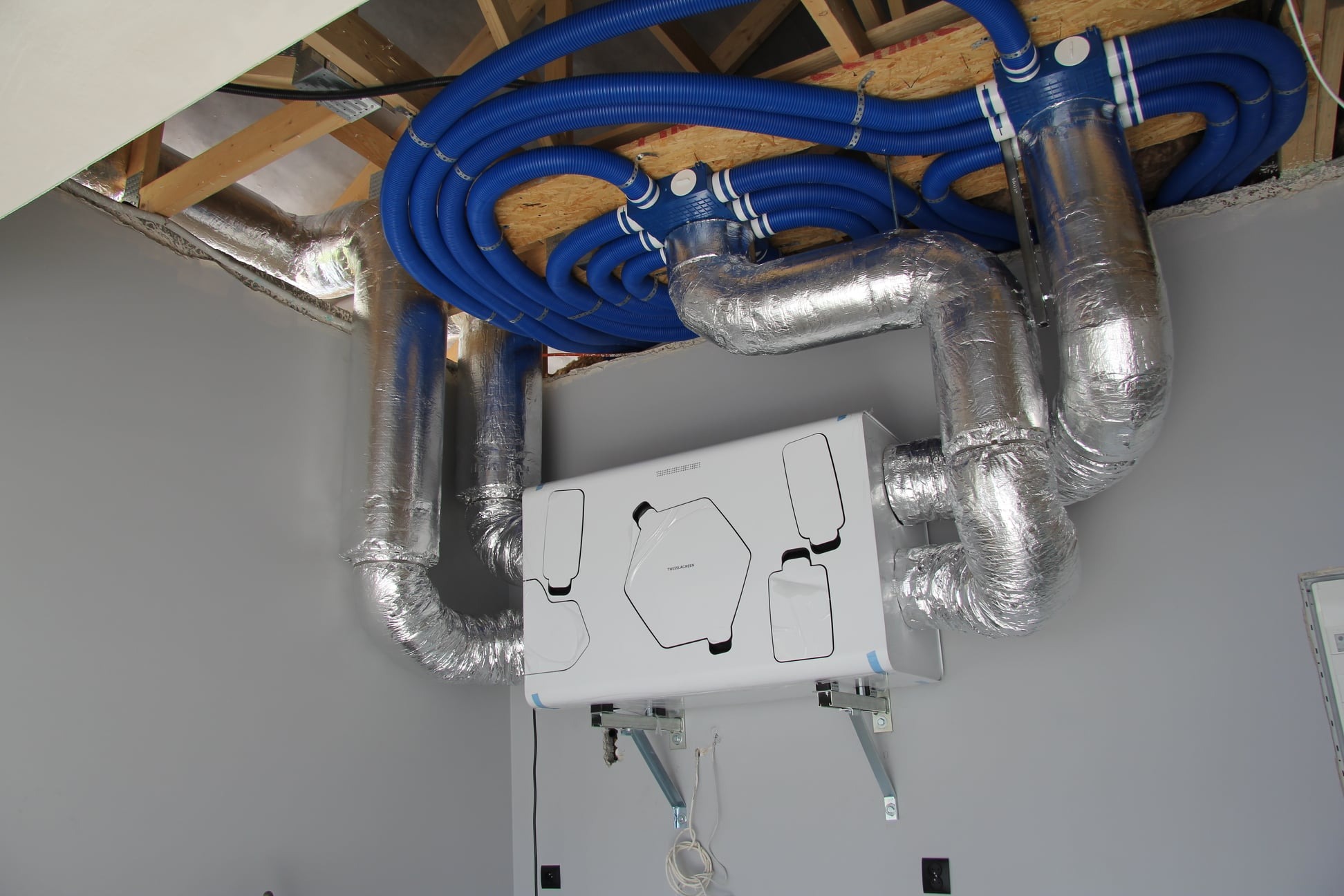 Centrala wentylacyjna z rekuperacją, widoczne niebieskie rury doprowadzające powietrze do rozdzielacza w suficie podwieszanym, srebrne przewody izolowane termicznie, biała obudowa urządzenia...