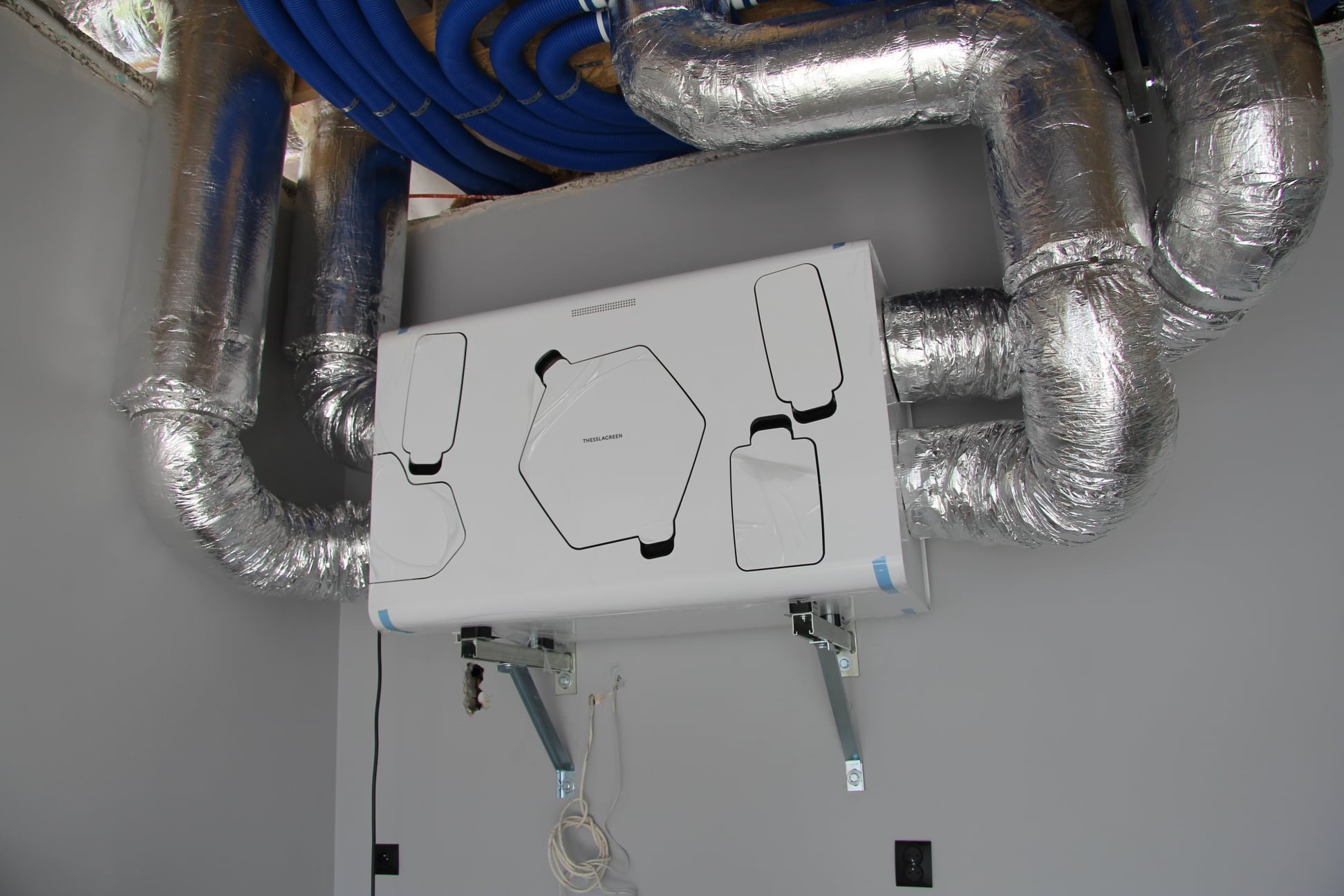 Zamontowany rekuperator marki Thessla Green z podłączonymi przewodami wentylacyjnymi w izolacji aluminiowej, widoczne niebieskie rury instalacyjne pod sufitem.