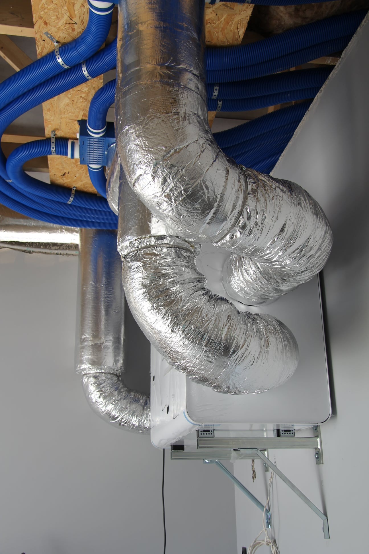 Rekuperator z podłączonymi przewodami wentylacyjnymi w izolacji aluminiowej i niebieskimi rurami doprowadzającymi powietrze, zamontowany na ścianie w pomieszczeniu w stanie surowym.