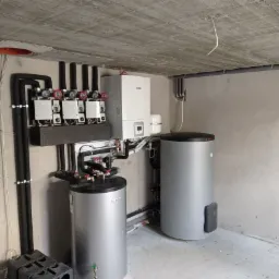 Kompletna instalacja grzewcza w surowym pomieszczeniu z betonowym sufitem, widoczne dwa srebrne zbiorniki, biały piec, rozdzielacz z pięcioma pompami obiegowymi oraz czarne rury.