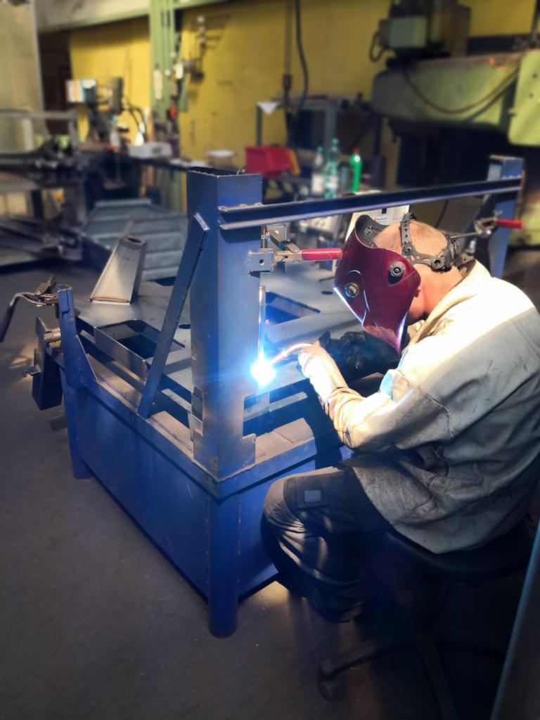 Pracownik w masce spawalniczej łączy metalowe elementy konstrukcji za pomocą spawarki, iskry i intensywne światło podczas pracy, warsztatowe tło z maszynami.