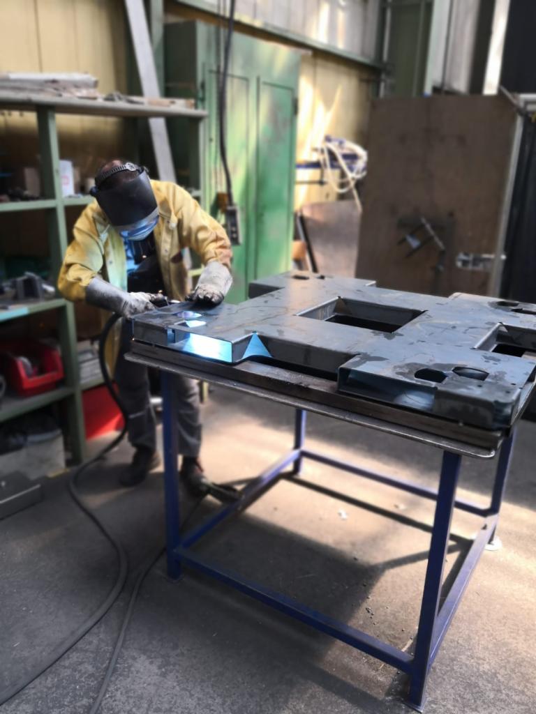 Spawacz w żółtej kurtce spawa metalową konstrukcję na niebieskim stole warsztatowym, iskry i dym widoczne w powietrzu.